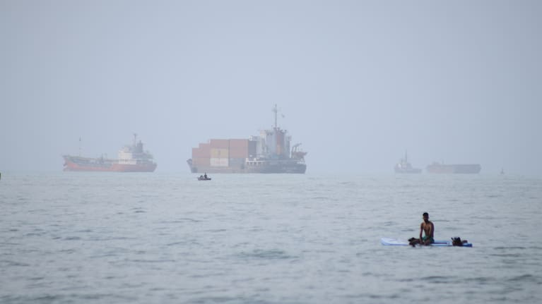 A man sits on a surfboard as a container ship and other commercial vessels appear anchored in the Strait of Hormuz off Bandar Abbas, Iran, Monday, July 27, 2026
