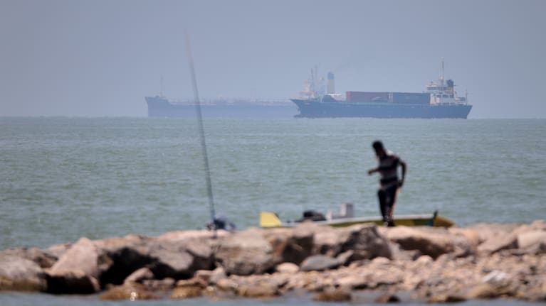 A man stands beside a fishing pole along the shore as cargo ships and commercial vessels are seen in the Strait of Hormuz off Bandar Abbas, Iran.