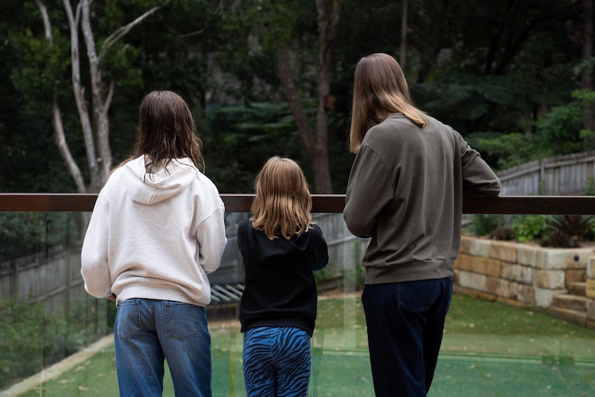 A woman and her two daughters standing at a pool fence looking at their backyard
