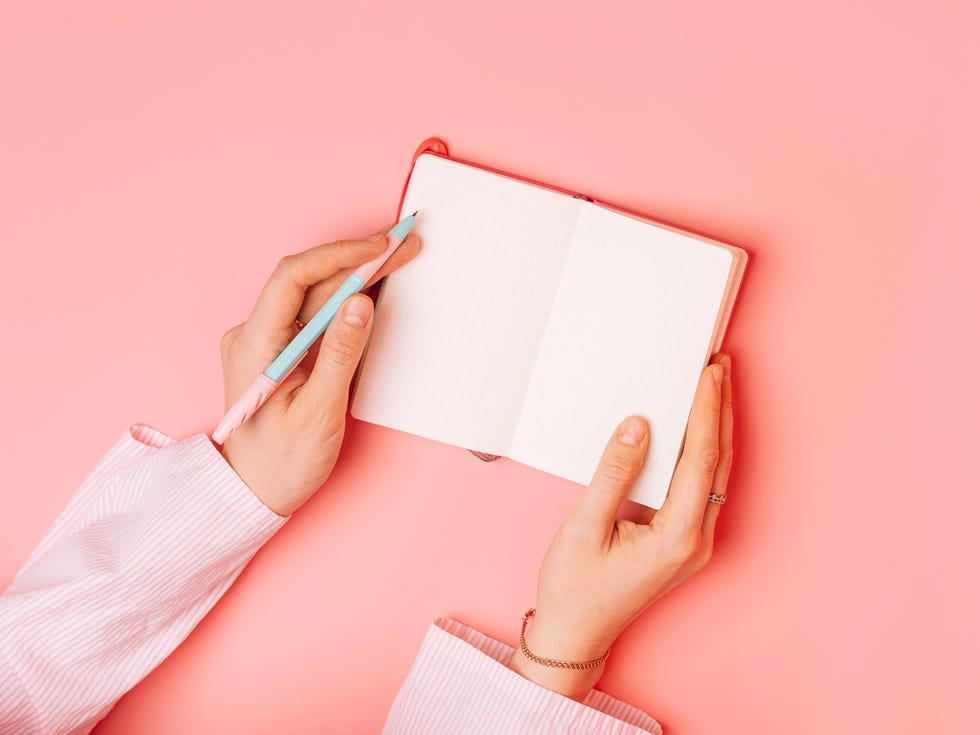 hands holding a pen and open notebook on a pink background.