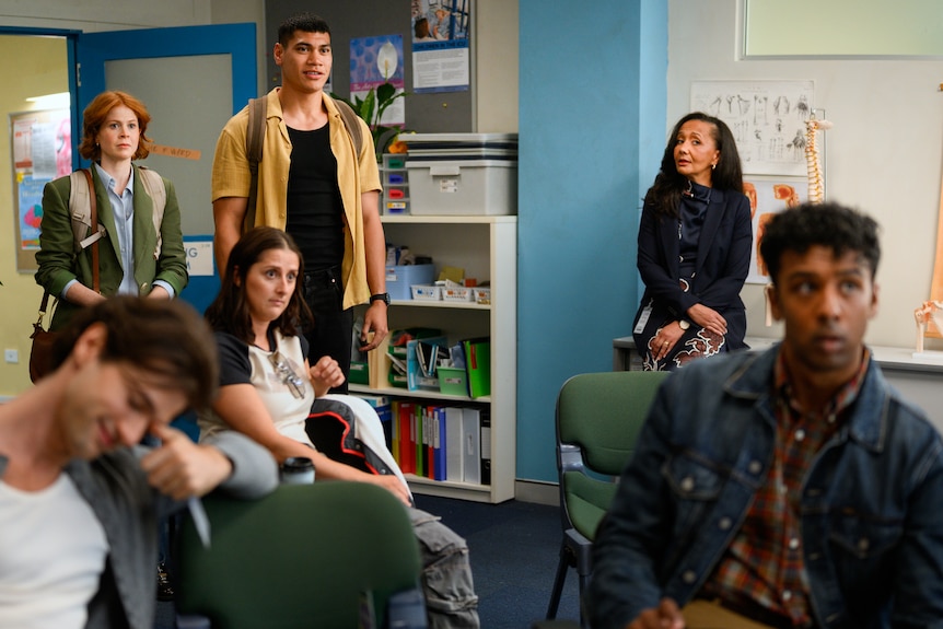 A TV still of a group of young people of different backgrounds, in casual dress, with a middle-aged woman, gather for a meeting.