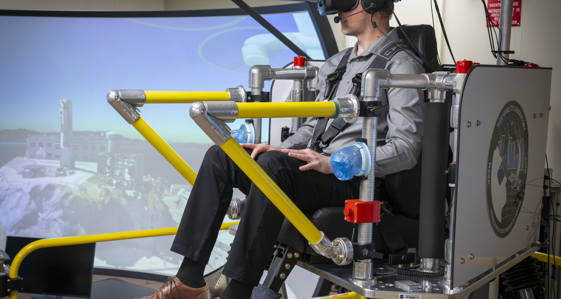 A brown and yellow platform holding a seat that is raised up in the air is holding one passenger. The passenger is wearing virtual reality goggles and a harness. There is a TV monitor to the right of the monitor showing the words “liftoff” and a second screen behind the passenger.