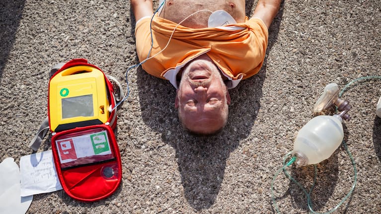 An unconscious man lying on asphalt during a medical emergency, connected to a portable defibrillator and oxygen mask. (file image)