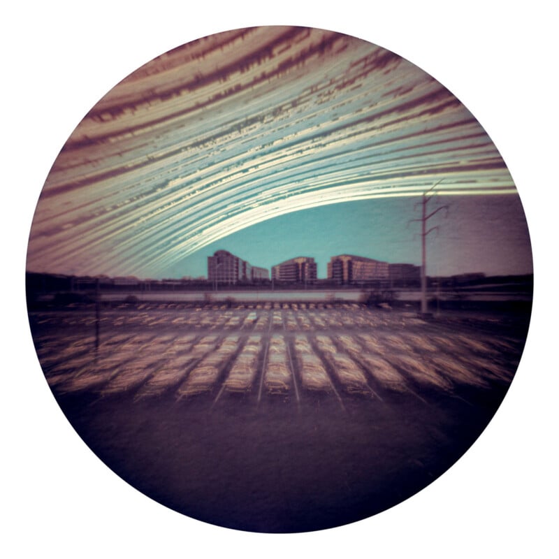 A circular photo shows a cityscape with streaks of light arcing across the sky above buildings and parked cars, captured with a long-exposure technique, creating surreal, sweeping patterns.