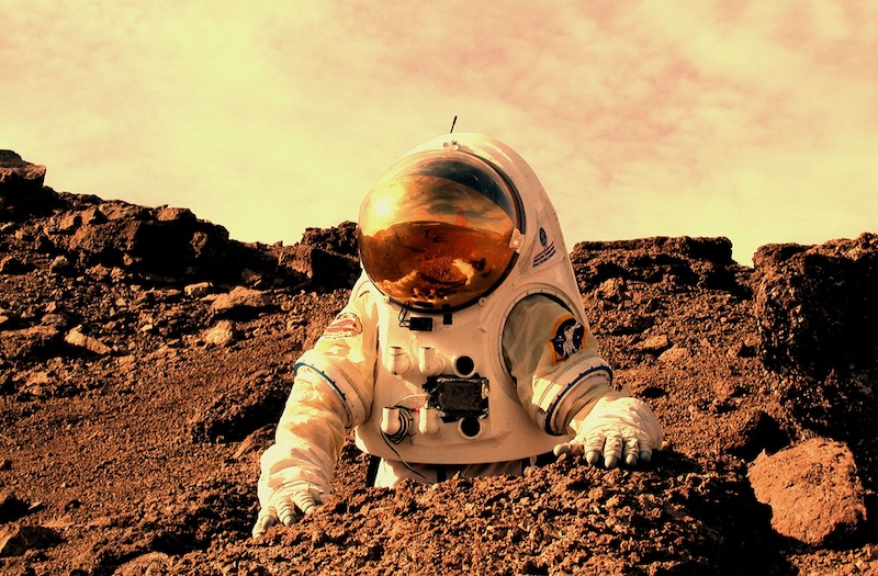 Microbes on Mars: Astronaut in bulky white suit standing near a big red boulder in reddish terrain with dusty yellow sky above.