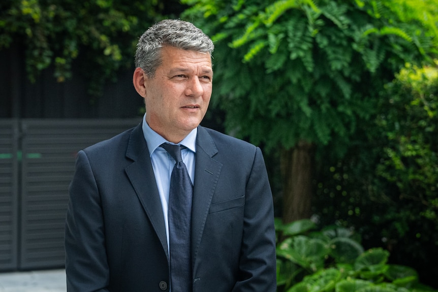 A grey-haired man in a dark suit stands in a garden.