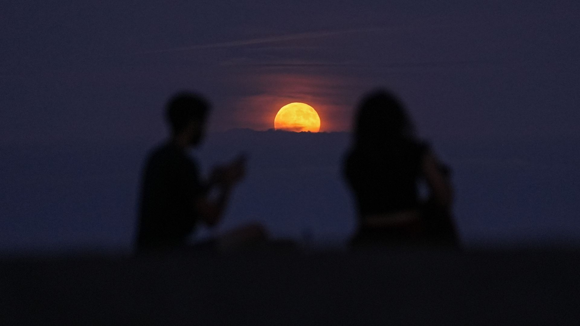 An orange full moon rises above a bank of clouds in a dark evening sky between the silhouettes of two onlookers.