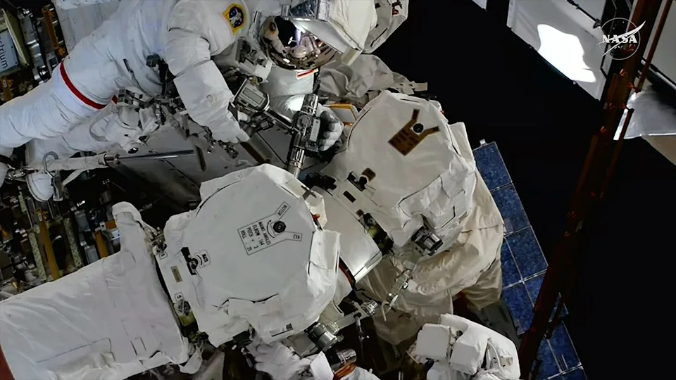Two astronauts in white spacesuits work to repair a robotic arm outside of a space station.
