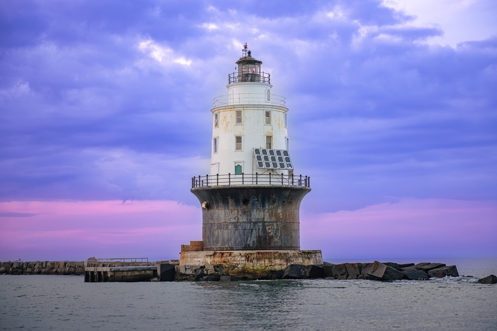 A lighthouse stands tall on the rocks against a purple-blue sky.