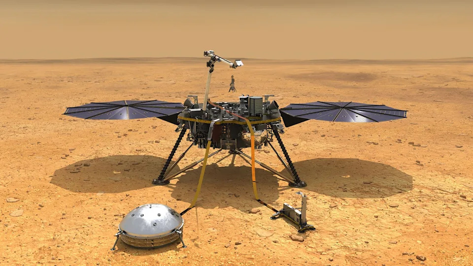a squat, round lander sits on three metal legs on a reddish-orange dusty surface, with two large wing-like solar arrays, one on each side