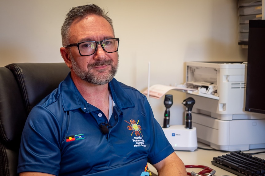 A man wears glasses and a blue polo shirt, sits in an office behind a desk with a computer and looks with neutral expression.