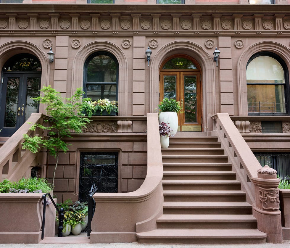 Brownstone building facade with steps, potted plants, and decorative details on the entrance and window.