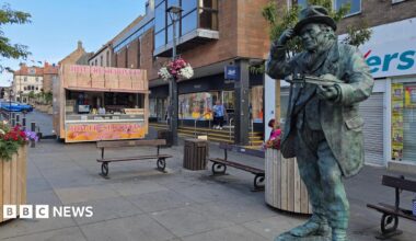 The bronze statue of Jimmy Stength is stood in the middle of a high street. A donut stall is stood near the benches to the left of the statue.