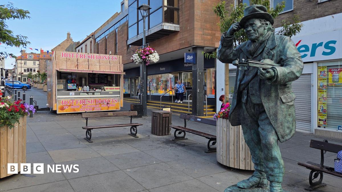 The bronze statue of Jimmy Stength is stood in the middle of a high street. A donut stall is stood near the benches to the left of the statue.