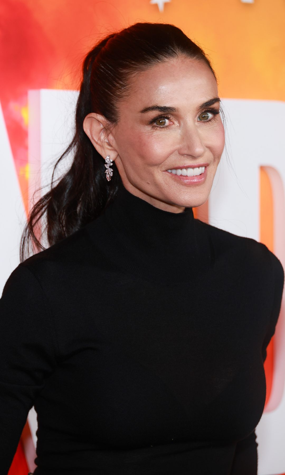 Demi Moore smiling, wearing a black turtleneck, and standing in front of a colorful backdrop.