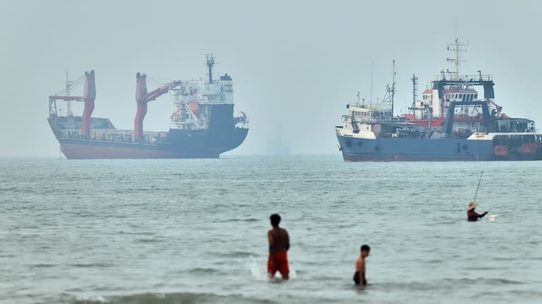 Children wade in the water with cargo ships at anchor in the background and a fisherman nearby, in the Strait of Hormuz off Bandar Abbas, Iran, Tuesday, June 30, 2026.