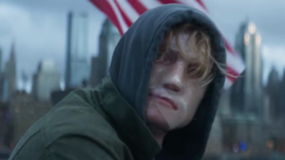 closeup of man in a dark hoody sitting outside, his disfigured face covered by a mask