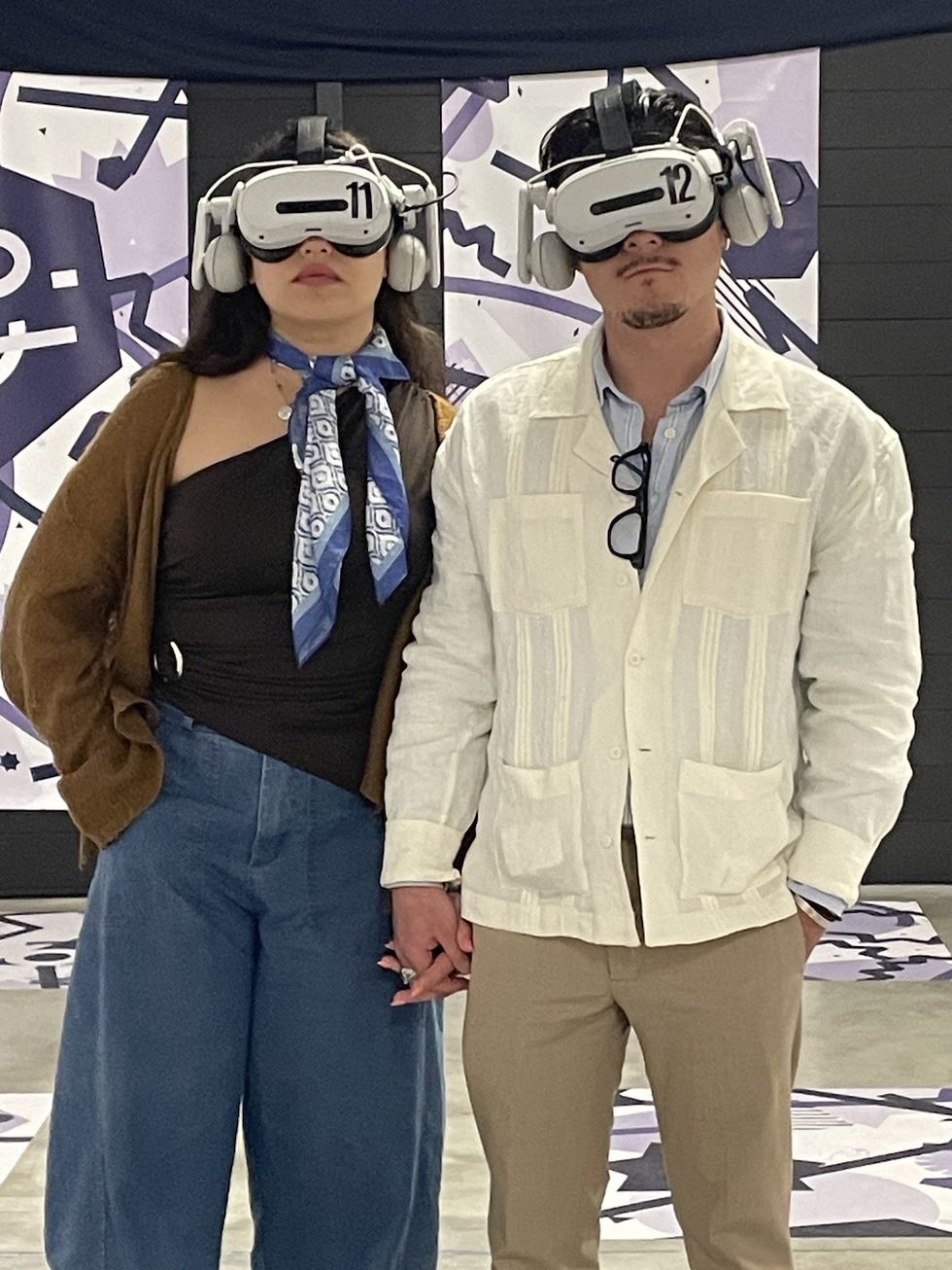 Couple holding hands wearing VR headsets in a modern exhibition space.