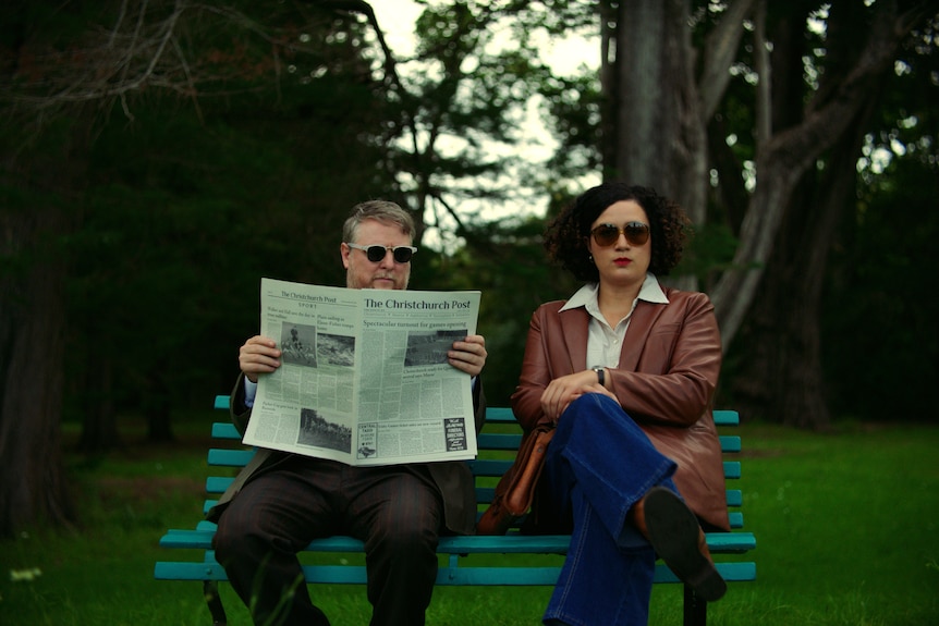 Tim Key sits on a park bench holding up a newspaper, next to Rose Matafeo in dark glasses