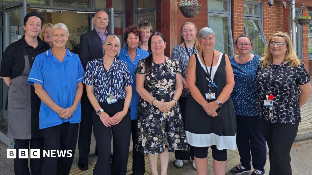 Staff members of the care home stand for a photo, they are smiling. Some wear nursing scrubs, while others wear office attire.
