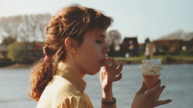 A colour photograph shows a young Nina outside di hospital, holding an ice cream cone with a large swirl of whipped cream in 1990. Nina dey in profile, with her hair tied back, and she wear a light-coloured top and a wristwatch. Trees and a few buildings dey visible for di softly blurred background across di water. Di image get di warm tones and candid feel of a family snapshot.