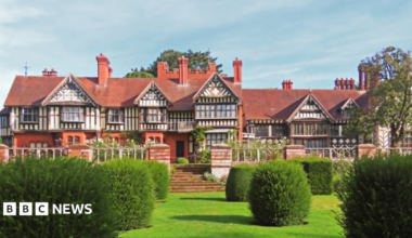 A large timber-framed building with tall brick chimneys and an ornamental garden with large bushes on a grass lawn