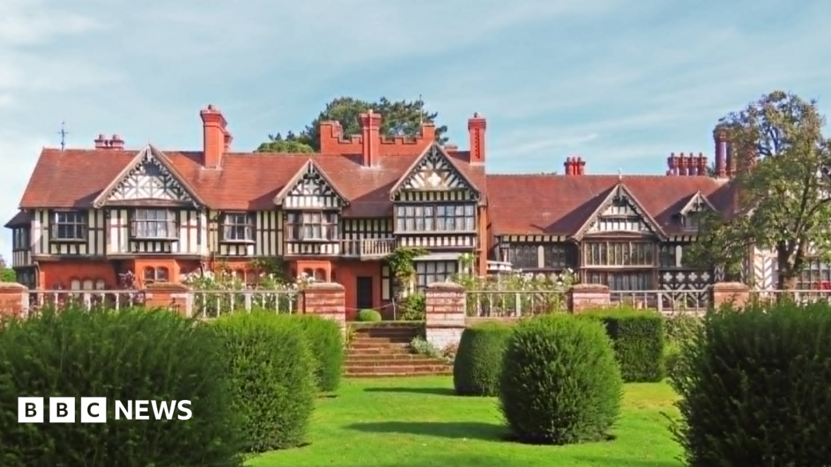 A large timber-framed building with tall brick chimneys and an ornamental garden with large bushes on a grass lawn