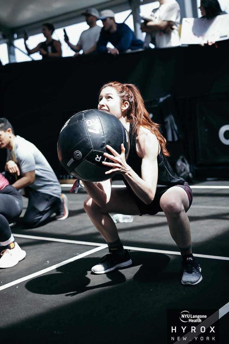 A woman performing a squat while holding a medicine ball during a fitness competition, with spectators in the background. A woman performing a squat while holding a medicine ball during a fitness competition, with spectators in the background.