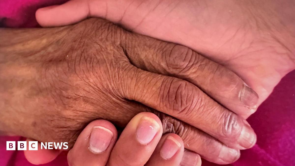 A close up of two people holding hands over a pink piece of fabric. The hand on the left is more wrinkly, that the one underneath, on the right.