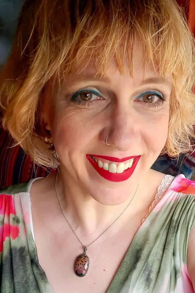 A present day photo show an adult Nina looking and smiling directly at di camera. She get short, light-coloured hair and wear a patterned top in shades of green and pink, a pendant necklace with an oval-shaped stone, and a small silver nose ring. Di image dey tightly cropped around di head and shoulders, with a softly blurred indoor background. Bright colours and natural lighting give di photograph a clear, modern appearance.