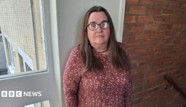 Pearl is standing in the stairwell of a block of flats looking to camera with an unhappy face. She has long brown hair and is wearing black glasses and a red dress with white floral pattern