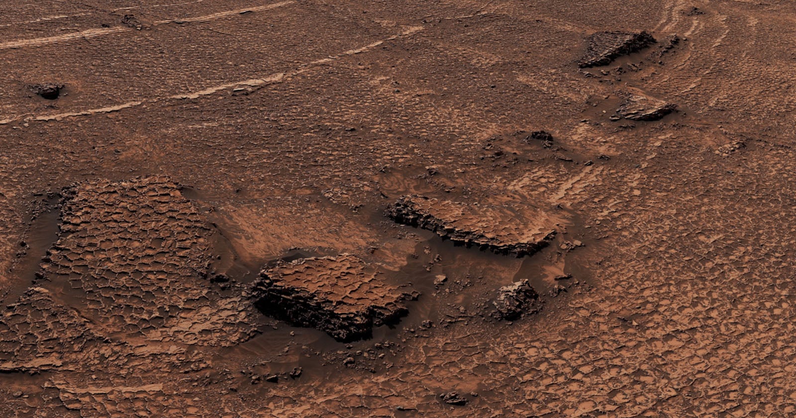 A high-angle view of a reddish, arid Martian landscape featuring polygonal patterned ground and several rocky outcrops.