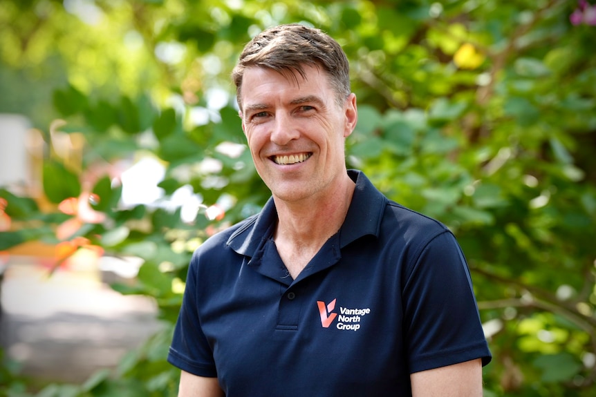 A smiling, dark-haired man in a polo shirt stands in front of some greenery.