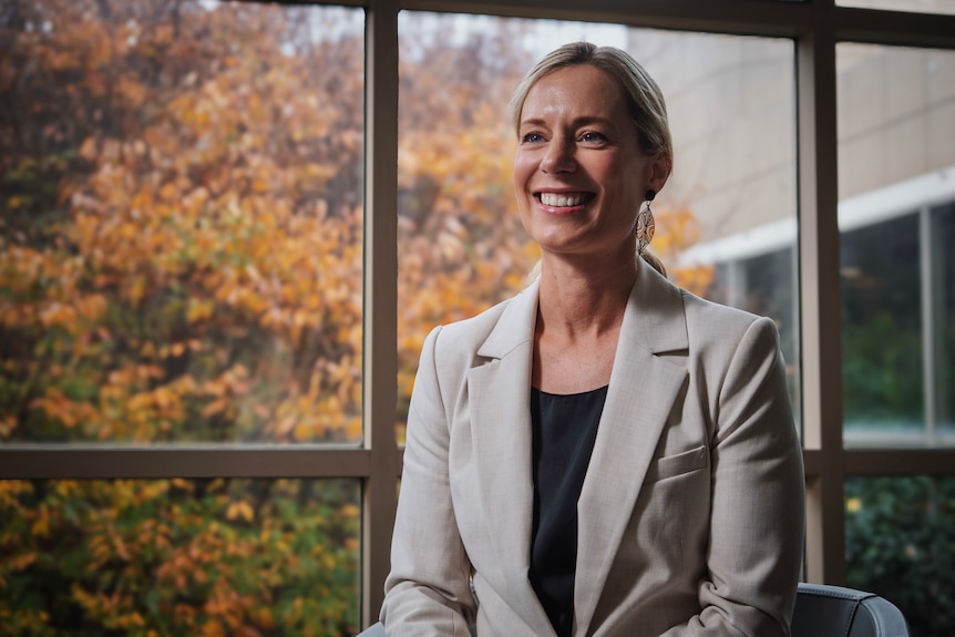 A woman smiles at the camera with a large window behind her
