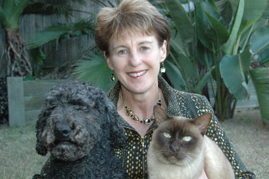 Jacquie with a dog and a cat, smiles, has short hair, large banana trees behind.