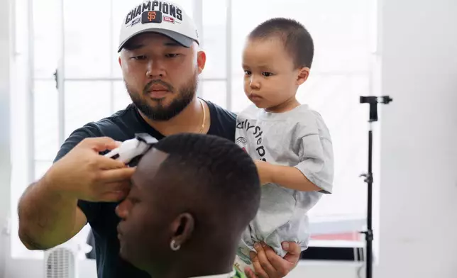 Jack Chang cuts Isaac Obamehinti's hair while holding his 2-year-old son Cairo Miu at his studio on Monday, July 13, 2026, in San Francisco. (AP Photo/Juliana Yamada)
