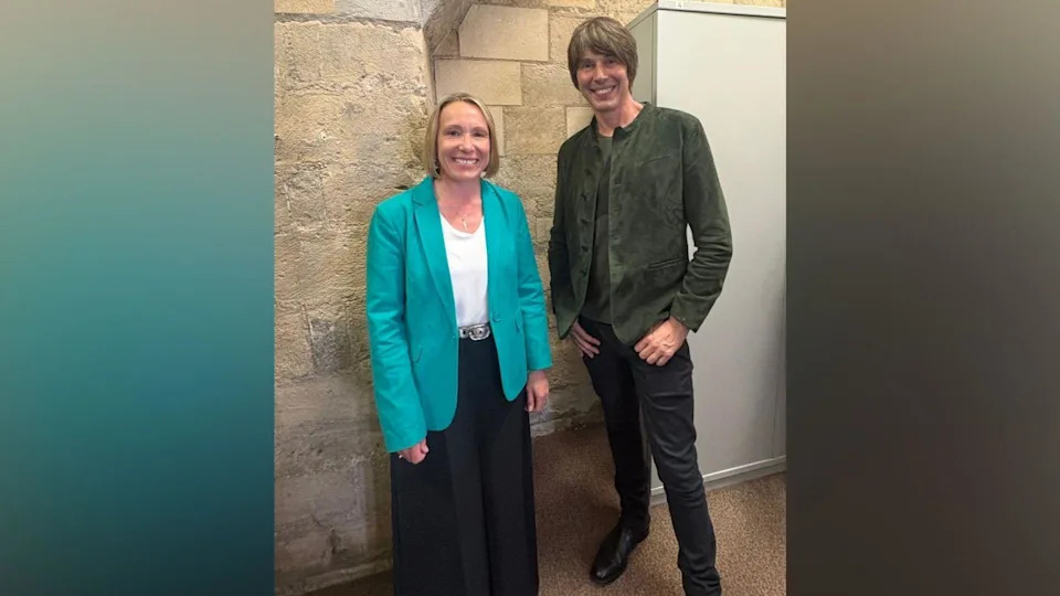 On the left, there is a woman in a turquoise-teal blazer, white t-shirt and black trousers. She has blonde ear-length hair and is smiling. On her right, there is a man with black jeans, black trousers and a dark grey jacket. He also has brown hair brushed to his left.