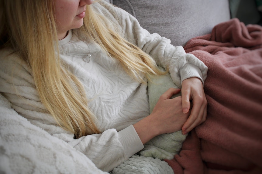 A woman with long blonde hair in a white jumper lies under a pink blanket with her hands resting on her stomach.