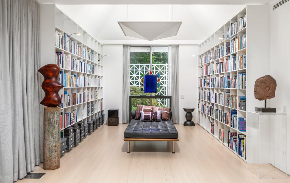 A modern library with shelves full of books, a sleek sofa, decorative sculptures, and large windows.