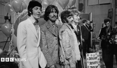 The Beatles post for a photo. Their clothing is from 1967. The photo is black and white. Some photographers are visible in the side of the image.