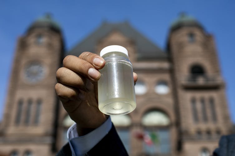 a hand holding a small bottle of polluted water in front of a large stone building