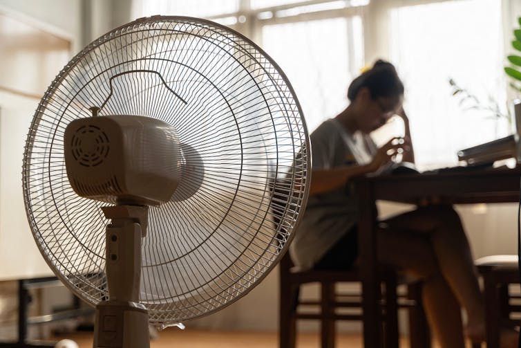 Woman sits at table at home with fan in foreground