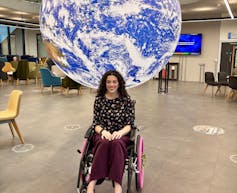 A brown haired young woman in a wheelchair with pink wheels and flowers smiles in front of a large illuminated globe suspended from the ceiling of Space Park Leicester, University of Leicester.