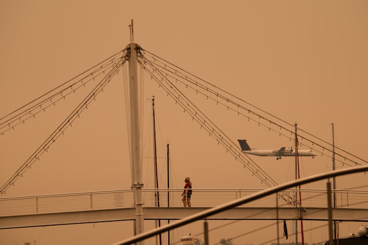 A plane in an orange-ish haze seen through the supports of a bridge.