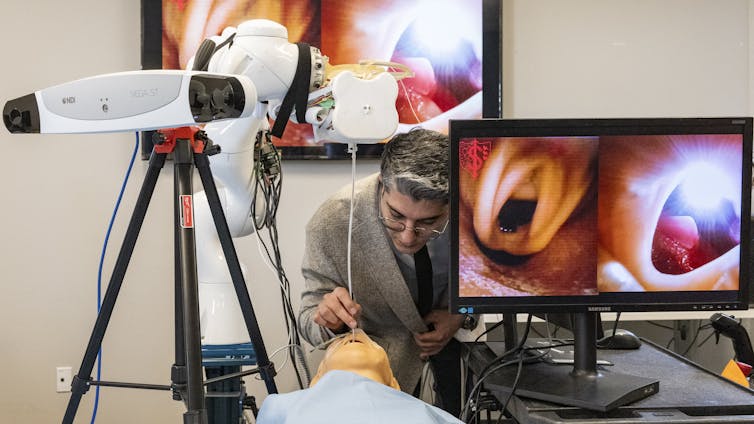 A man in glasses operates on a dummy patient in-between a robot and a large computer screen.