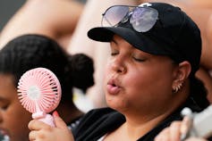 A woman outdoors in a baseball cap fans herself with a handheld fan