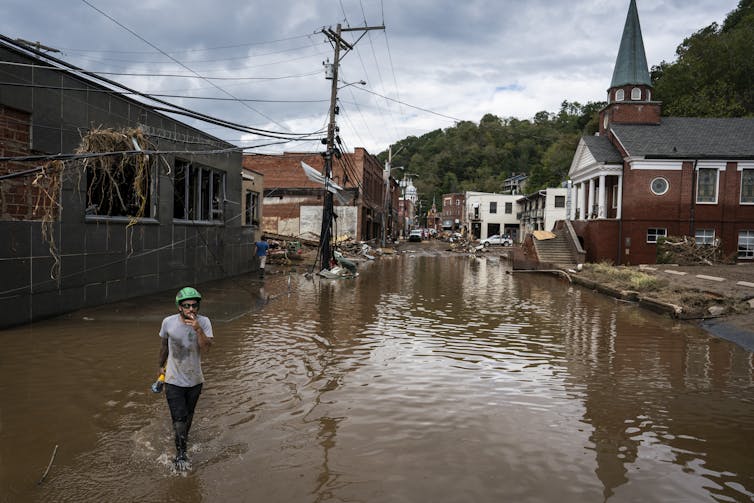 A person walks through a flooded street with damaged buildings, including a church, behind them.