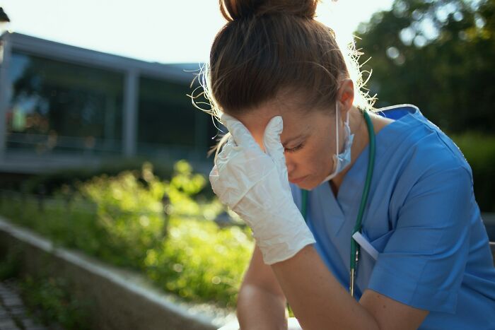An exhausted ER staff member in scrubs and gloves, holding her head, reflecting the stress of dealing with ridiculous patients.