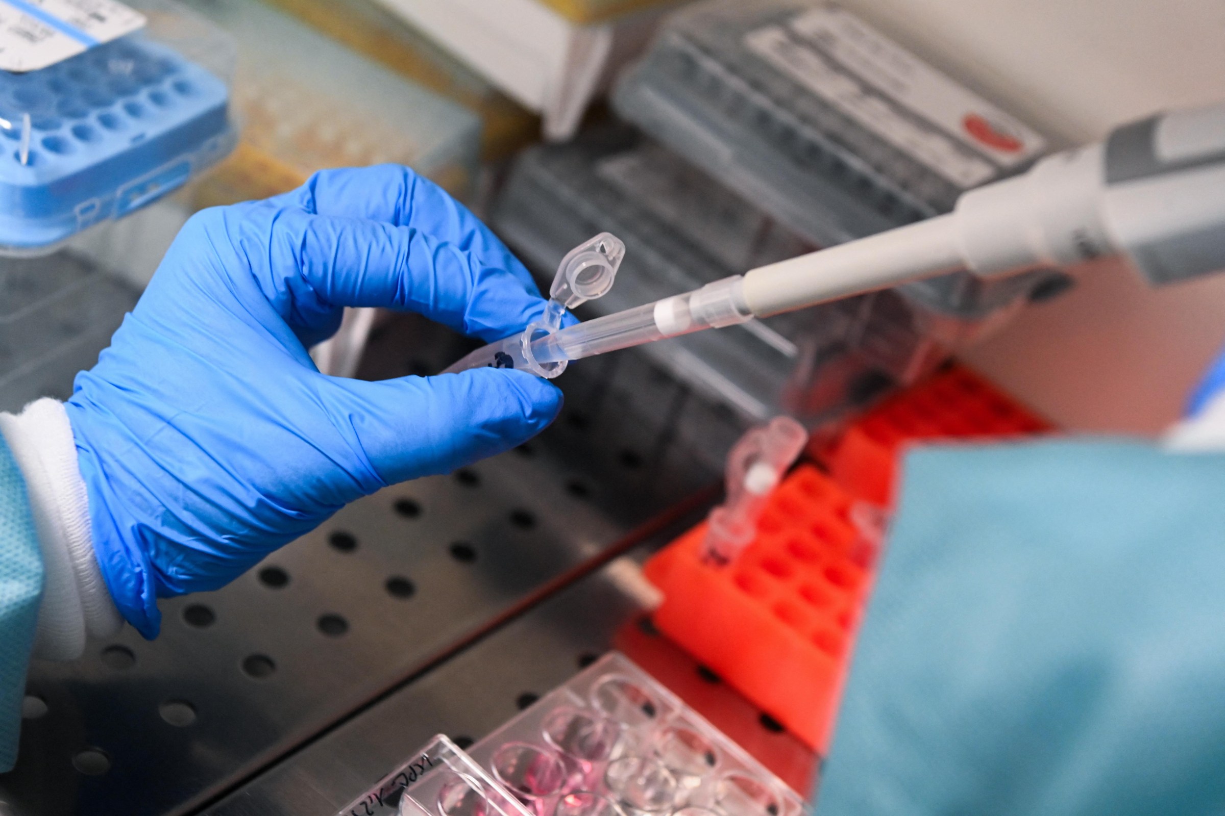 A blue-gloved hand holds a vial to a large pipette, transferring mRNA material.