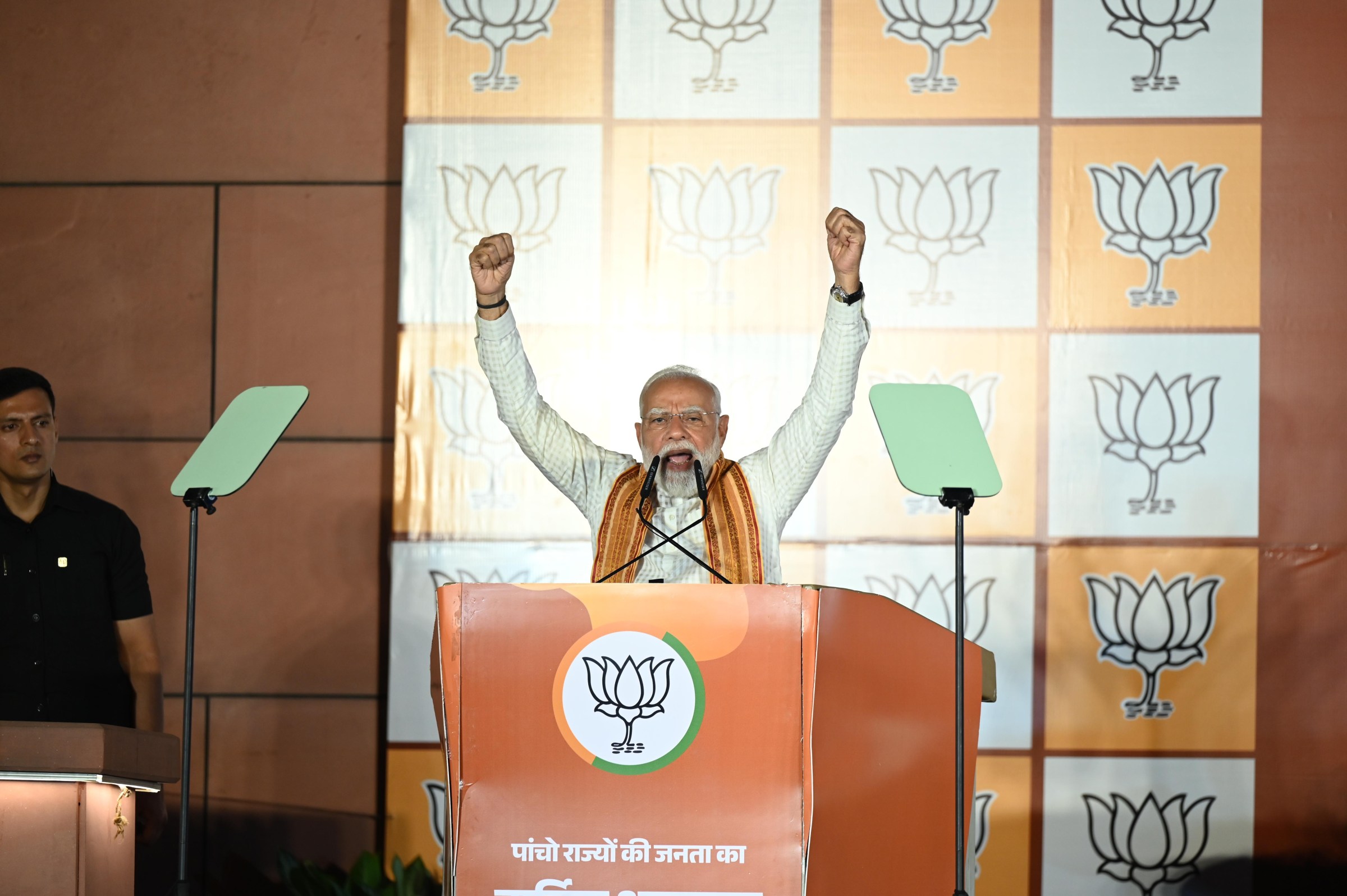 Narendra Modi raises both arms with fists over his head while speaking into microphones from a podium.